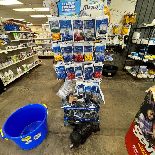 Display of agricultural spray tips in a store aisle with various colored packages on a stand. Nearby, a large blue bucket and a black tool rest on the floor.